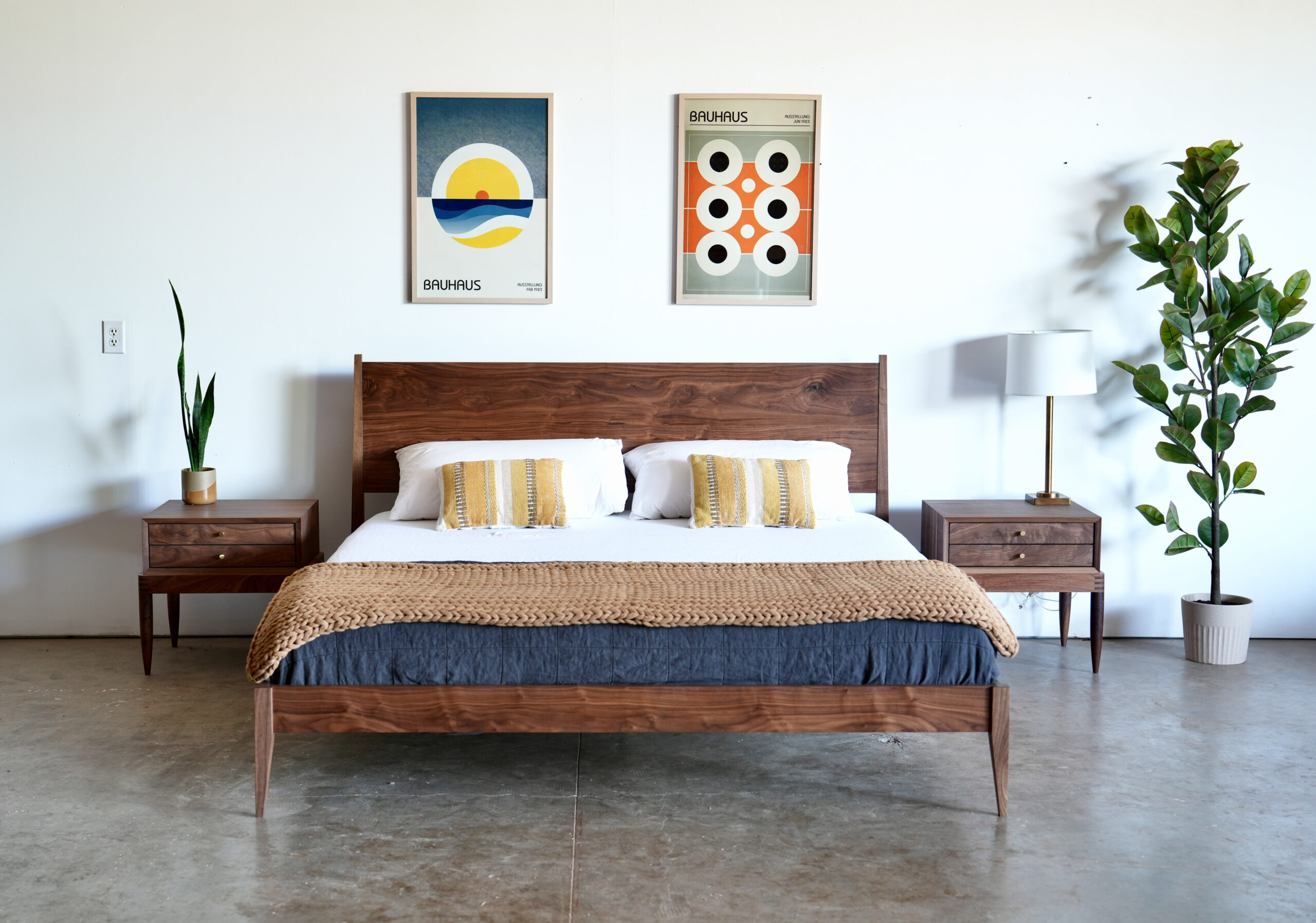 A walnut shaker style midcentury modern bed with tapered legs. A mattress, pillows, and a throw blankets are on the bed. There is a matching set of side tables next to the bed..