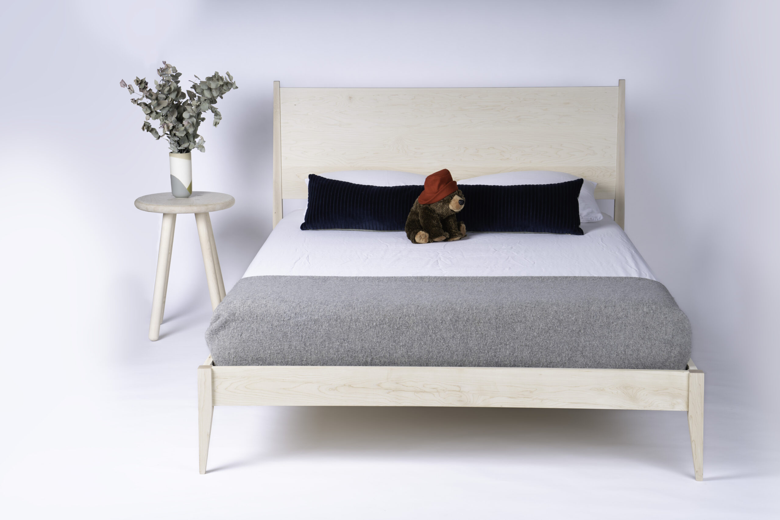 A solid maple bed with a teddy bear in a red hat on top of the mattress and a simple round side table with a plant.