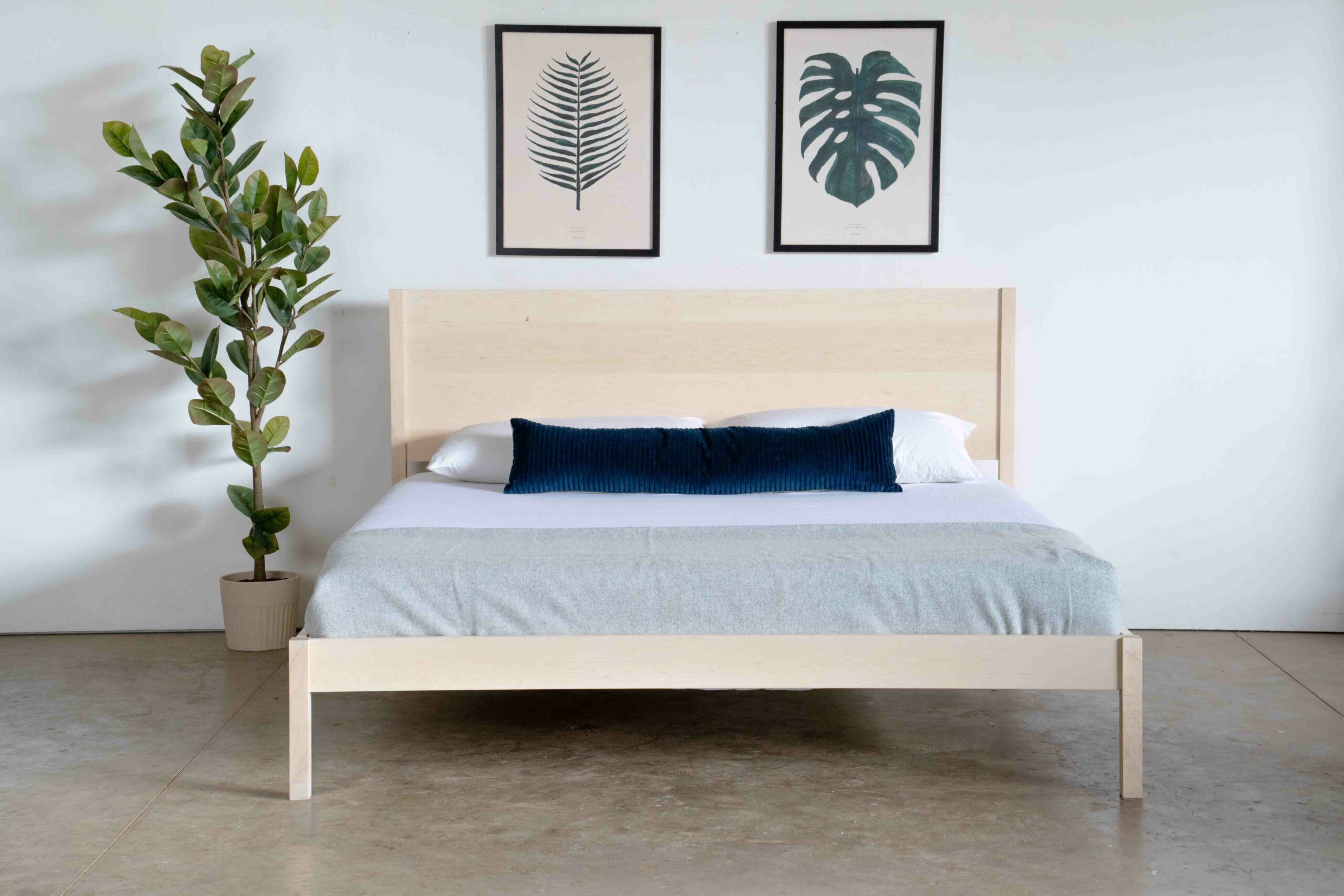 A simple platform bed in maple with a mattress and pillows, standing next to a plant.