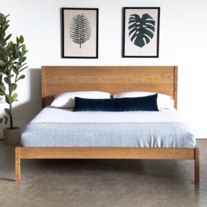 A simple platform bed made of cherry with a mattress and pillows next to a plant.