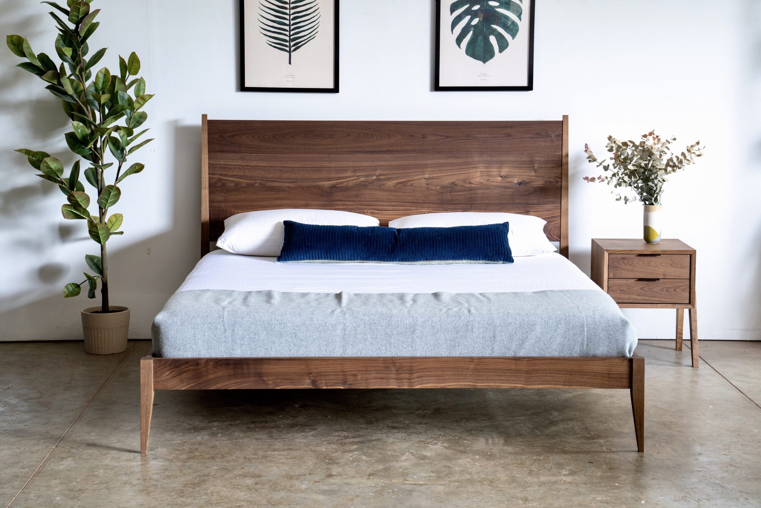 A simple walnut bed made up with a mattress and pillows. A two drawer walnut nightstands is next to it.