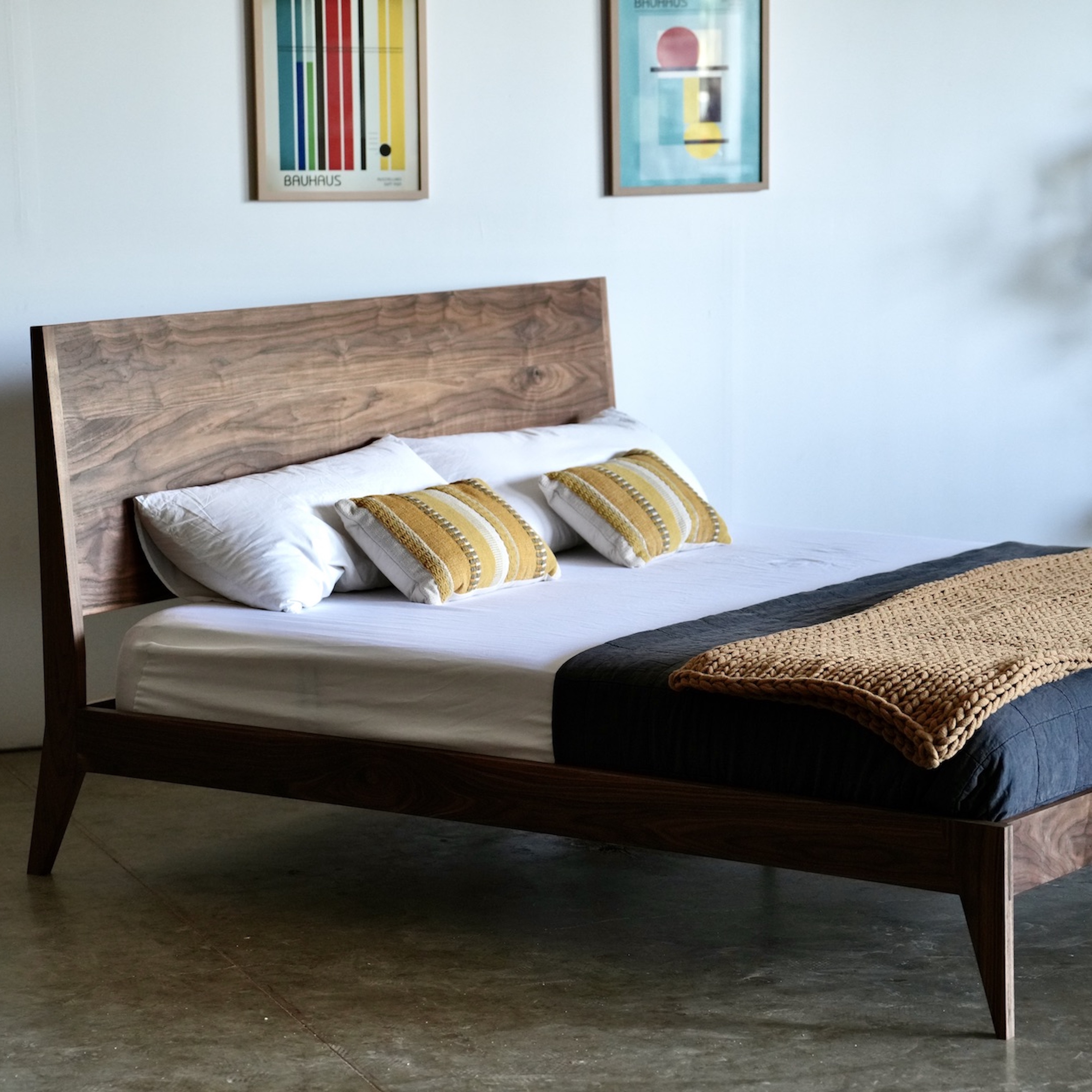 A solid wood bed with a slightly slanted headboard. It has white sheets and a yellow throw blanket and pillows.
