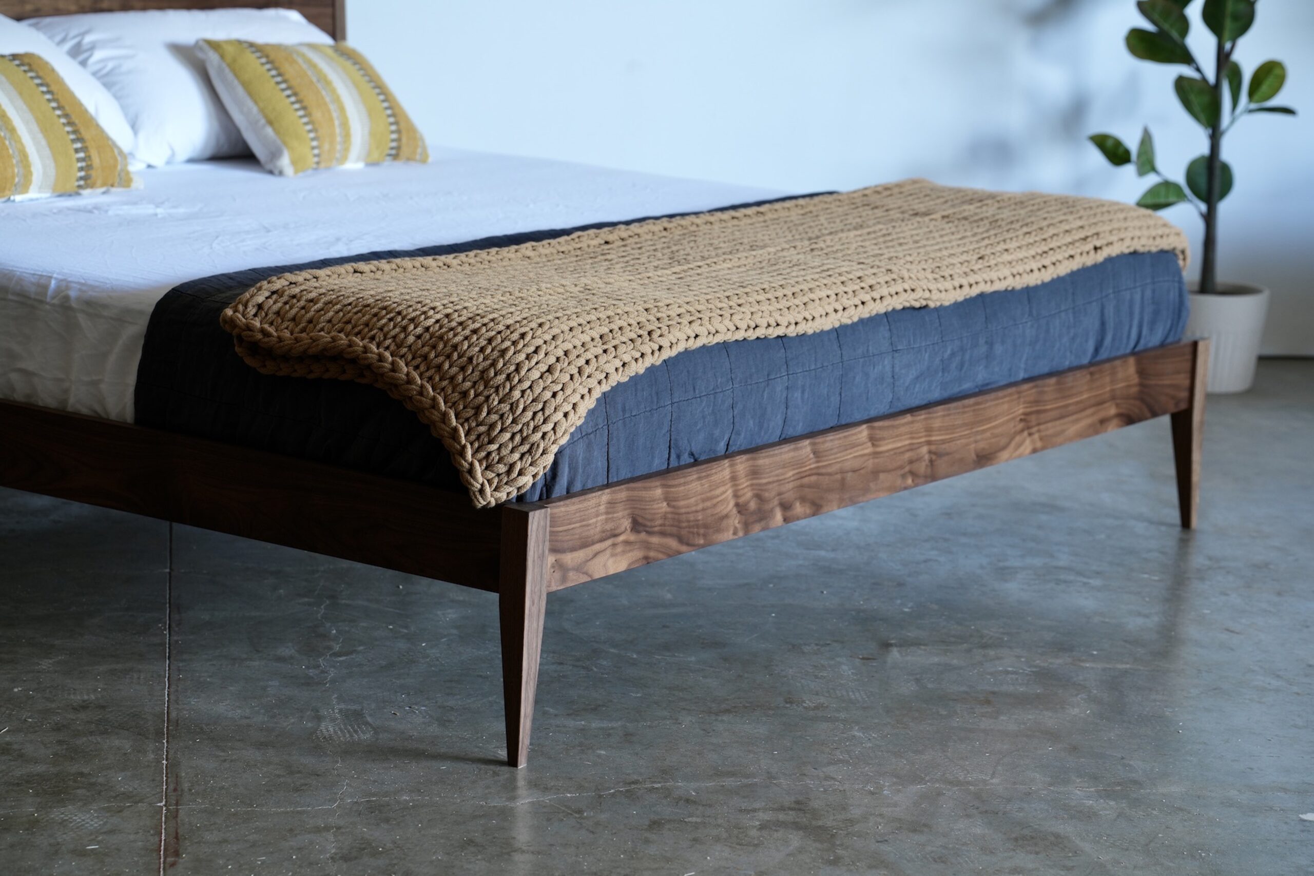 A close-up of the end of a walnut shaker style midcentury modern bed with tapered legs. A mattress, pillows, and a throw blankets are on the bed.
