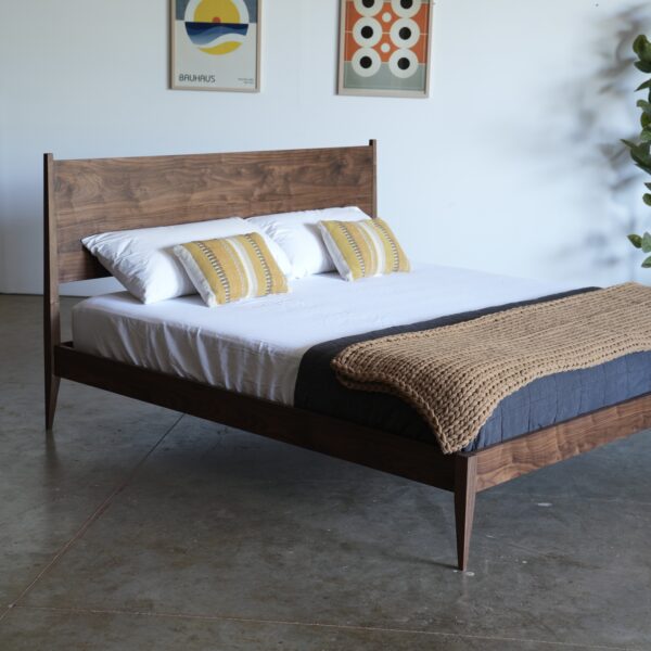 A walnut shaker style midcentury modern bed with tapered legs. A mattress, pillows, and a throw blankets are on the bed.