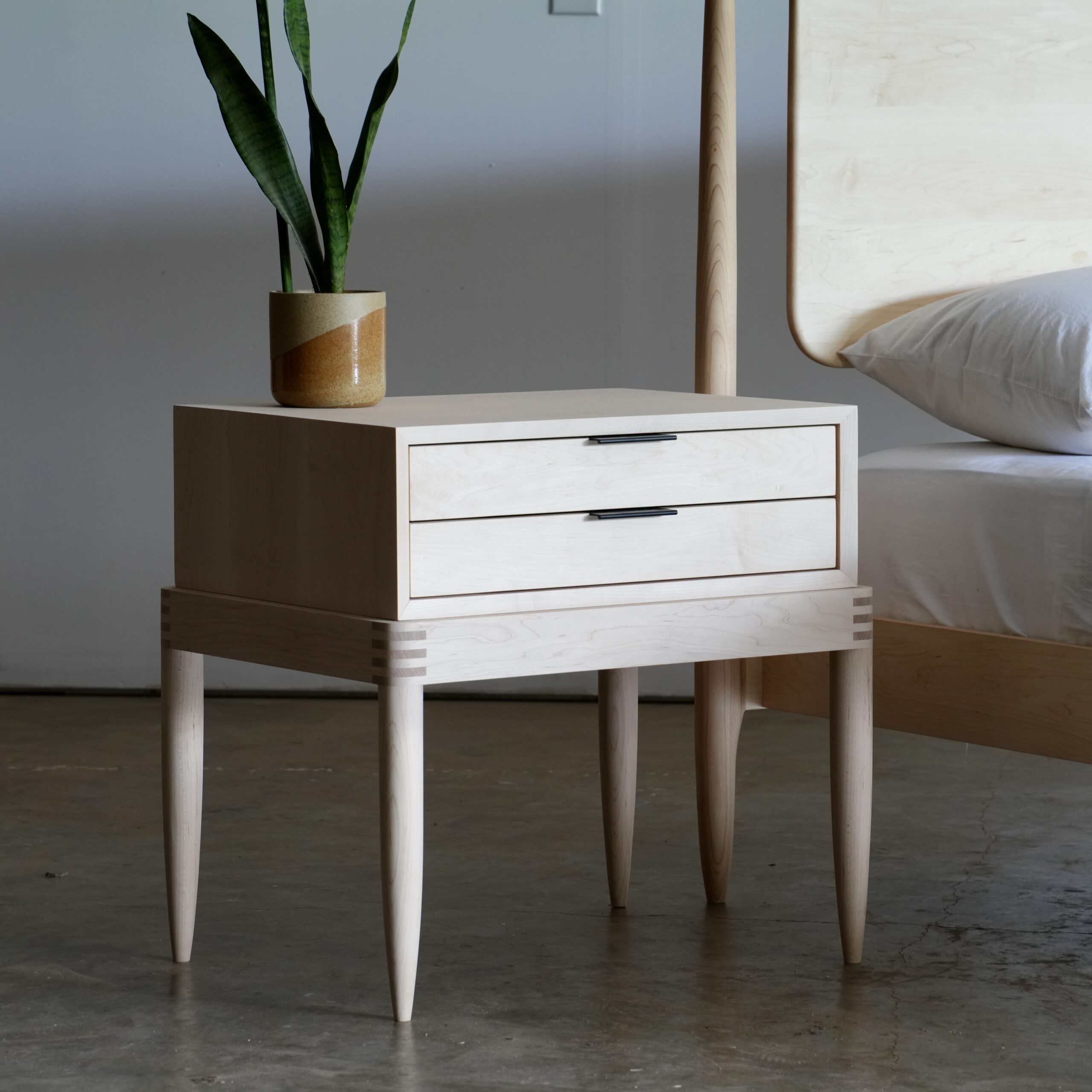 A simple two-drawer solid maple nightstand with exposed joinery at the base and black ledge pulls.