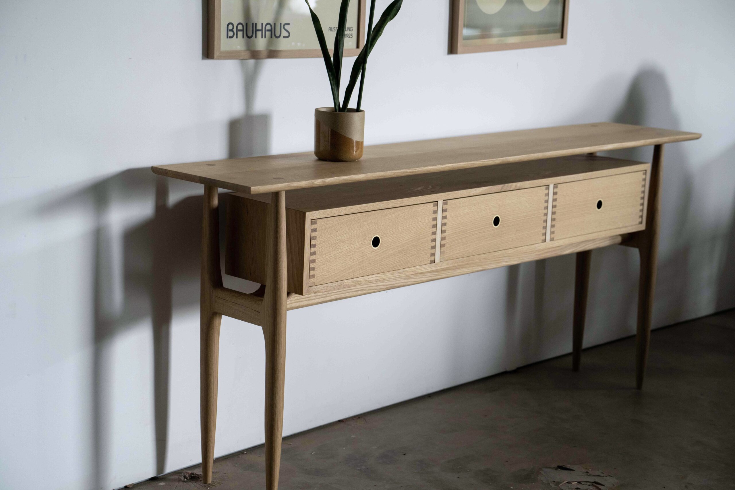 A narrow midcentury modern entry table made of white oak with three drawers.