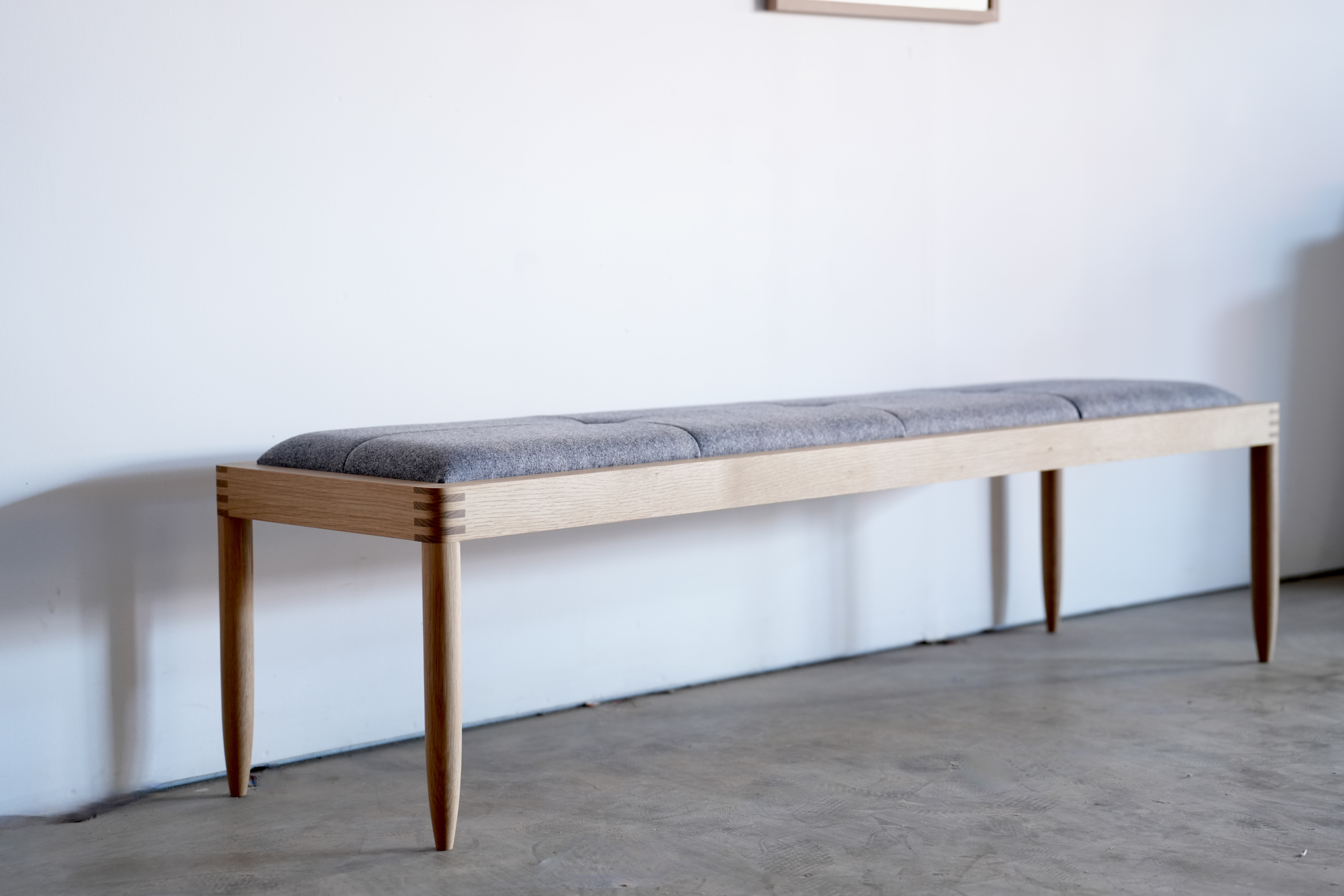 A simple white oak bench with exposed finger joints and an upholstered gray wool top.