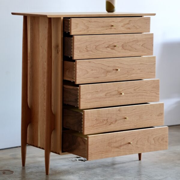 A 6 drawer cherry midcentury modern tallboy dresser with brass knobs in the center of each drawer.