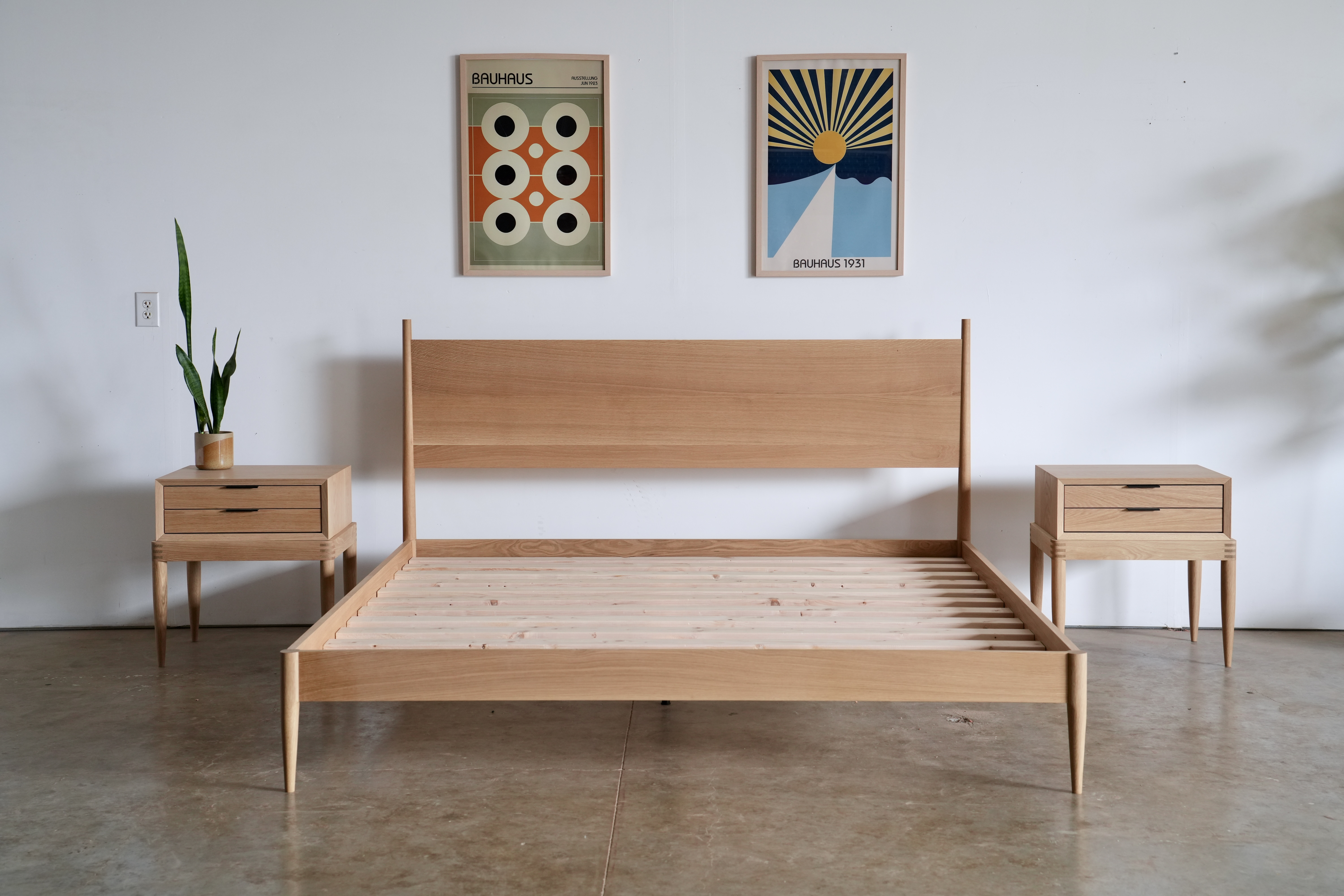 A simple midcentury modern bed made of white oak. There is no mattress so one can see the slats and two matching oak nightstands sit on either side of the bed.