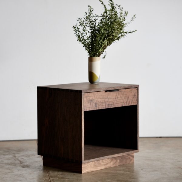 A simple solid wood nightstand with a single drawer and open box below.