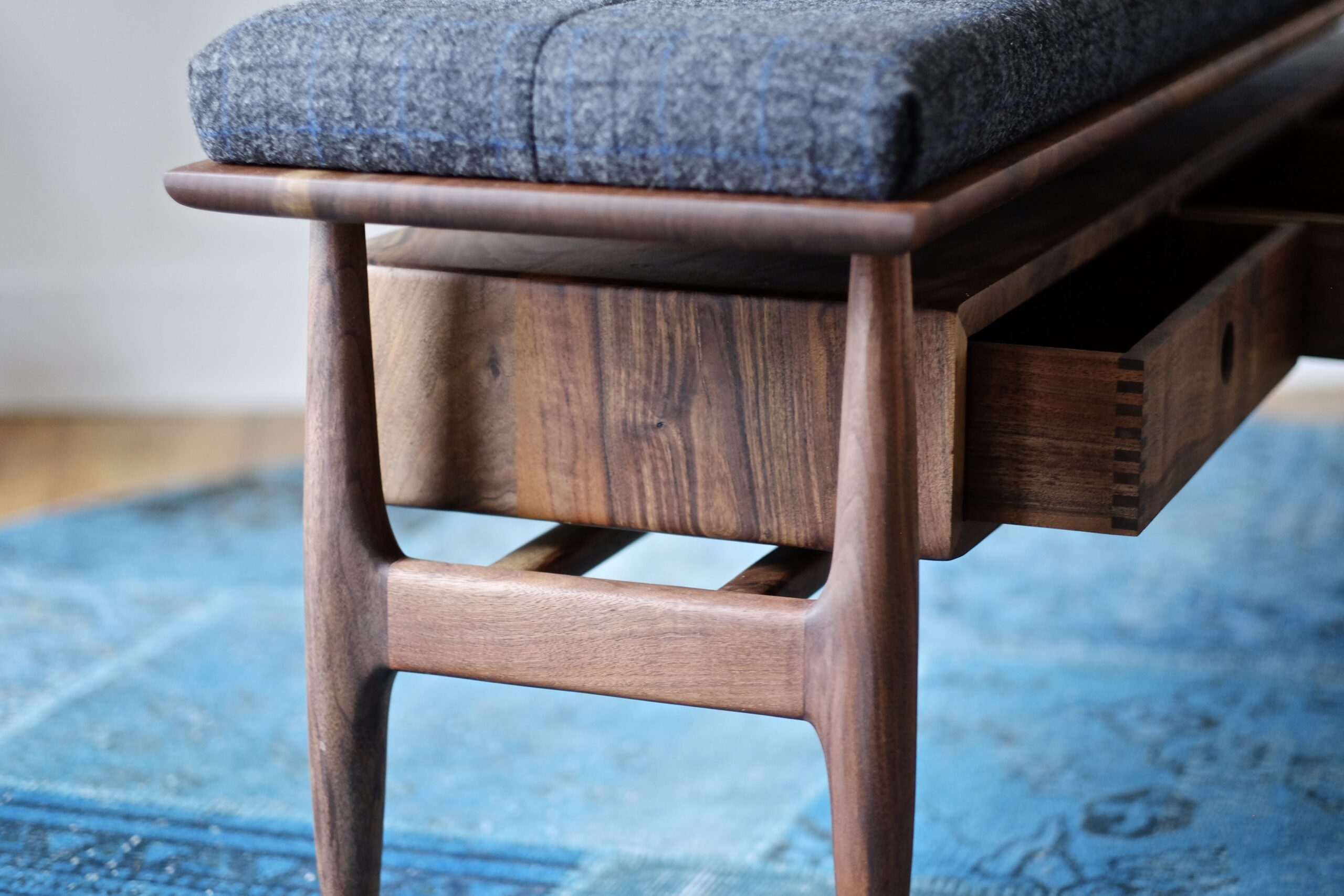 A walnut bench with a gray wool upholstered cushion and three drawers.