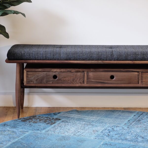 A walnut bench with a gray wool upholstered cushion and three drawers.