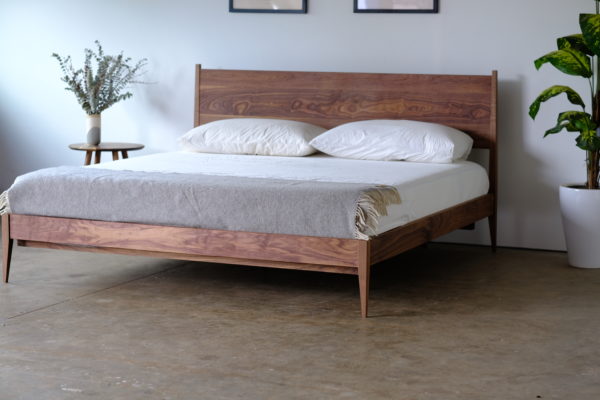 A walnut shaker style midcentury modern bed with tapered legs. A mattress, pillows, and a gray blanket are on the bed.