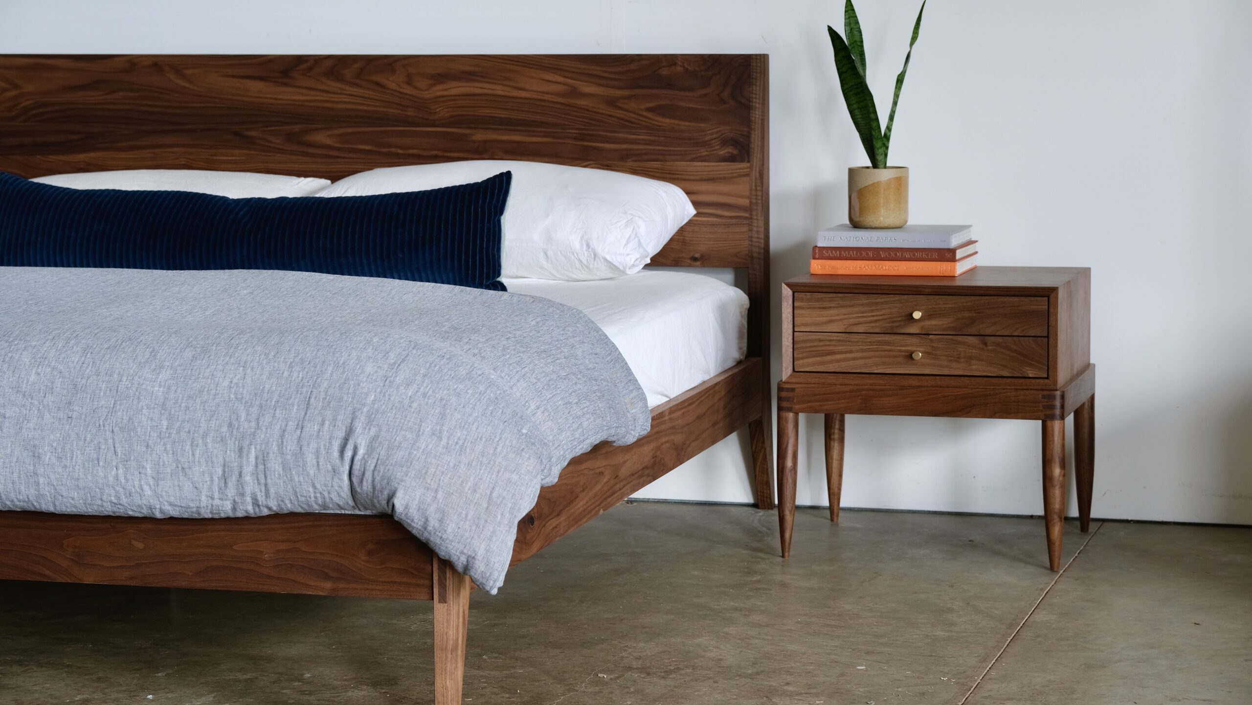 A walnut bed next to a solid walnut nightstand with books and a plant on top.