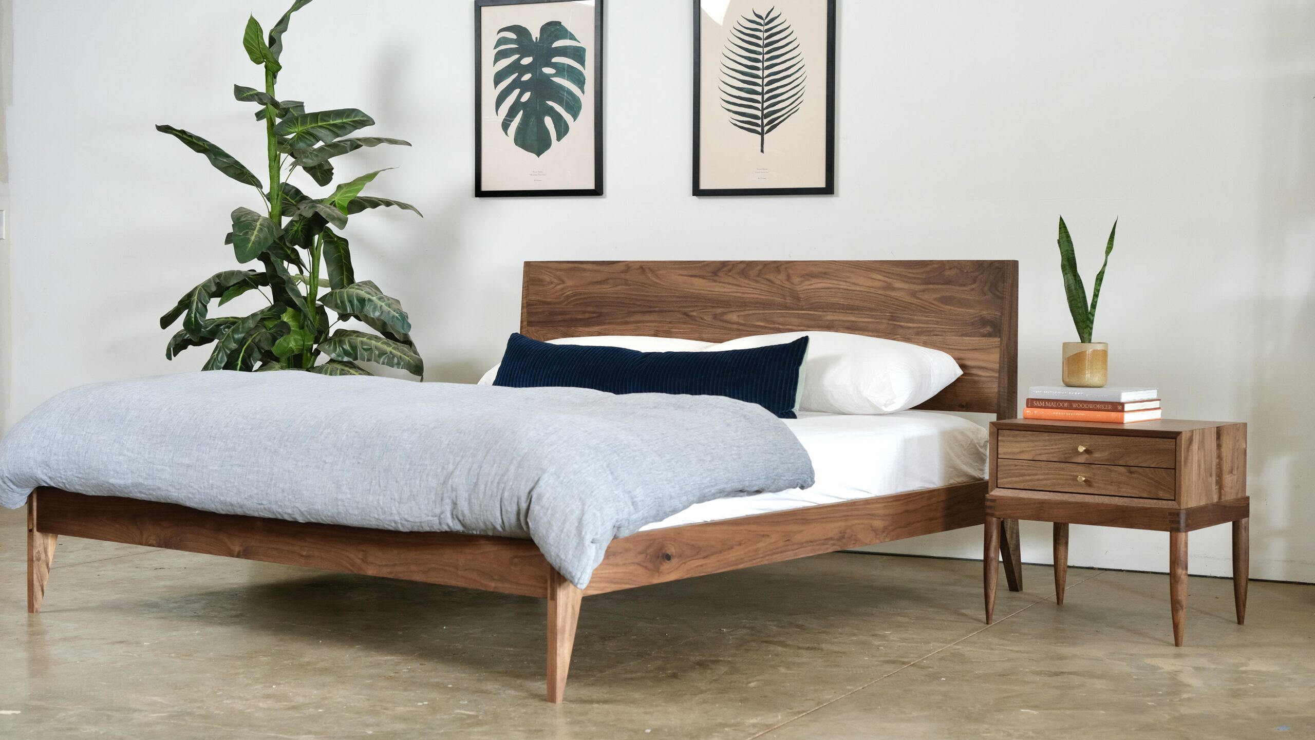 A solid wood bed next to a solid wood nightstand, both made of walnut, with books and a plant on top of the nightstand. Pictures are on the wall and a bigger plant on the other side of the bed.