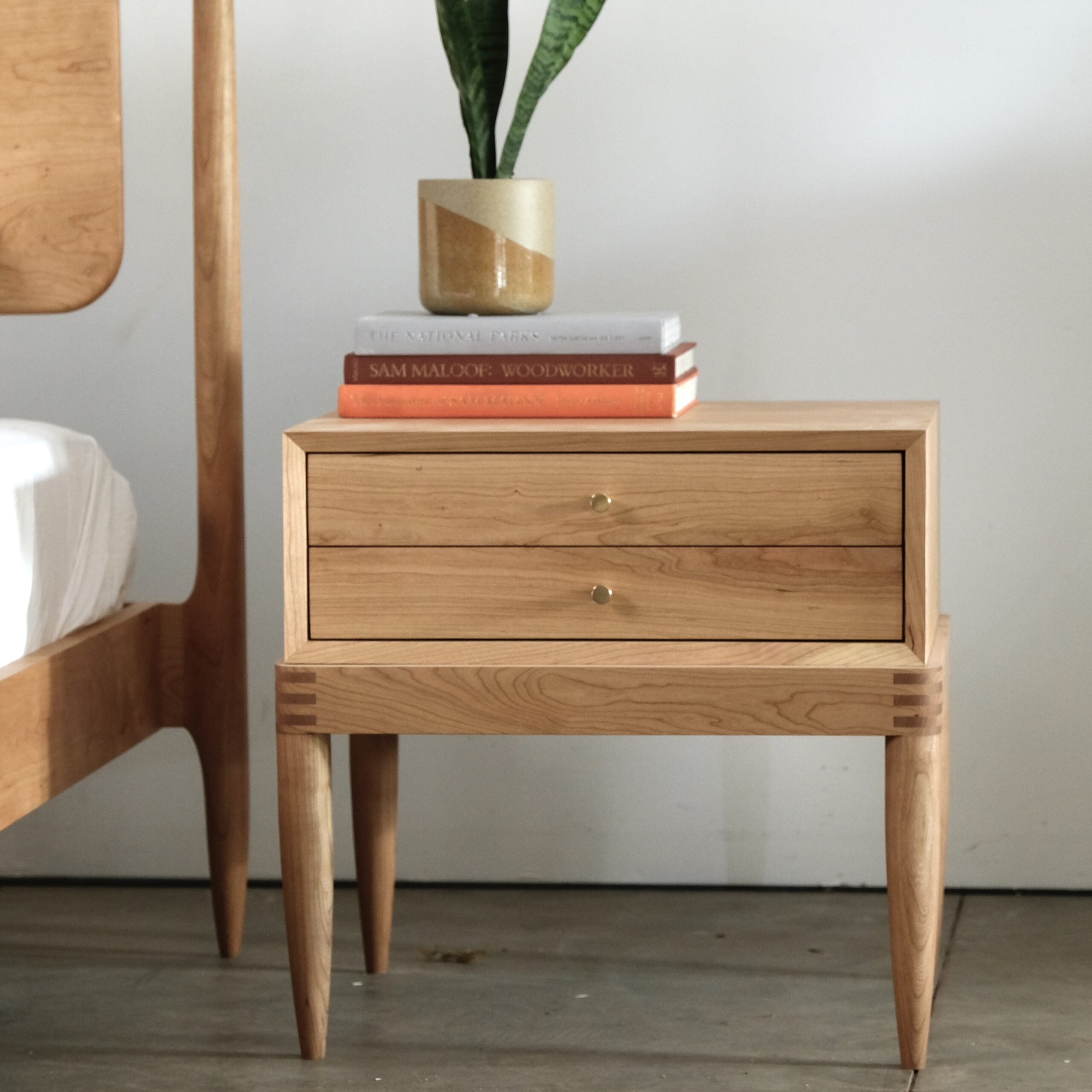 A simple two-drawer solid cherry nightstand with exposed joiner at the base and conical brass pulls.