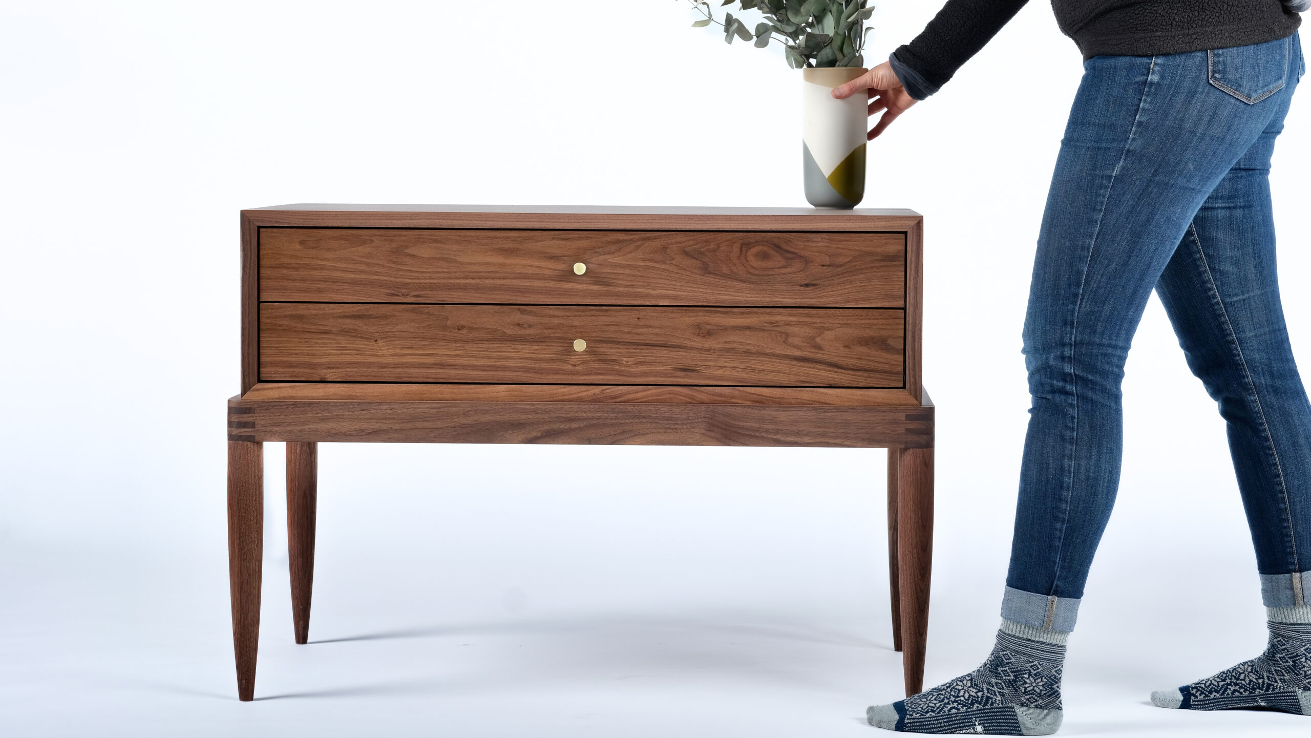 A solid wood nightstand made of walnut with two shallow drawers and round brass pulls. Someone is setting a vase on top.