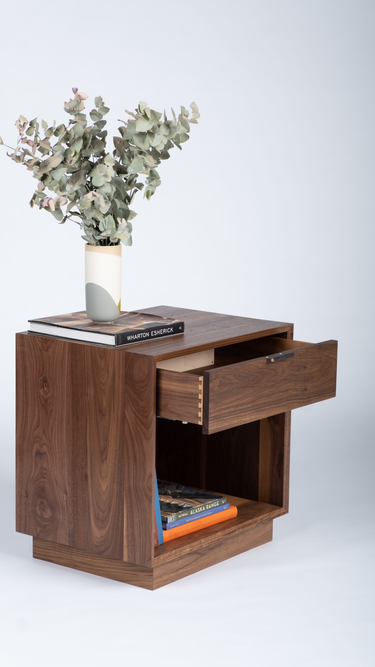 A box-shaped minimalist side table with an open space on the bottom holding books and one drawer. Made of solid walnut.