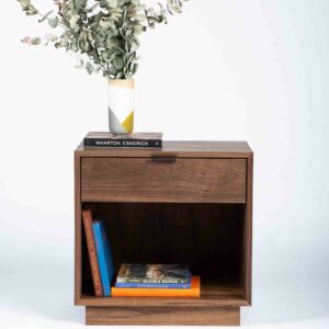 A box-shaped minimalist side table with an open space on the bottom holding books and one drawer. Made of solid walnut.