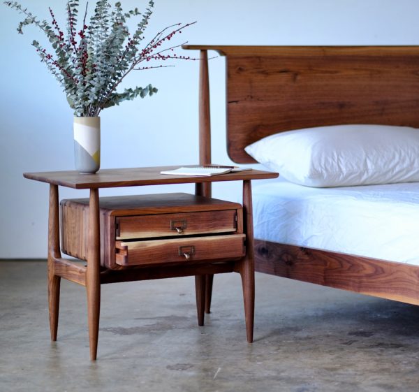 A handmade side table that is Scandinavian style, with the drawers appearing to float under the table top and made of solid walnut. Next to a walnut bed with a similar style headboard.