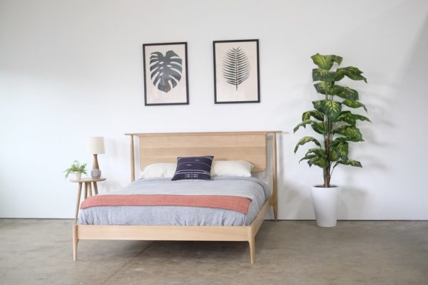 A mid century modern style oak bed with and oak nightstand and plant next to it.